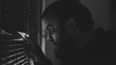 Ein Mann mit Bart späht mit dem Finger zwischen seiner Jalousie aus einem Fenster