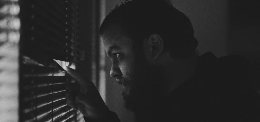 Ein Mann mit Bart späht mit dem Finger zwischen seiner Jalousie aus einem Fenster