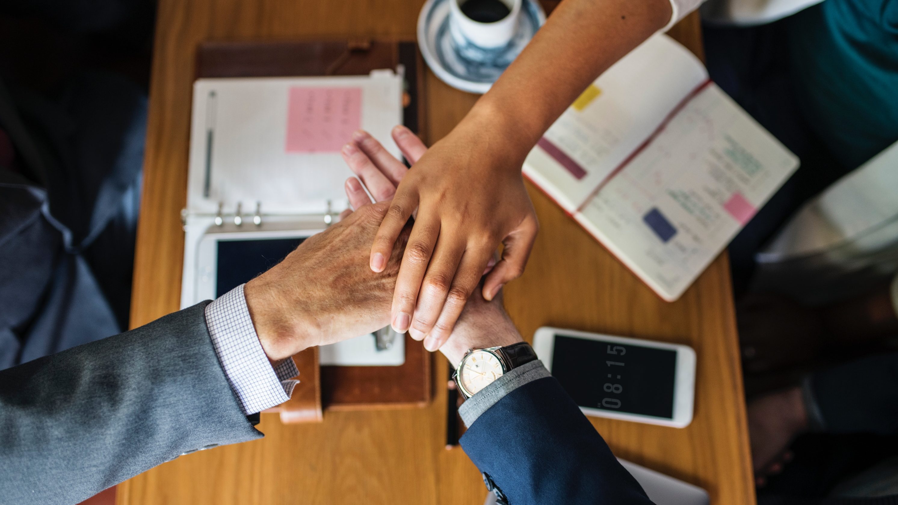 Über einem Tisch mit verschiedenen Schriebwaren und einem Handy, schlagen drei Hände zusammen ein.