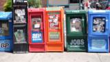 Aufgereihte Straßenzeitungsverkaufscontainer in unterschiedlichen Farben.