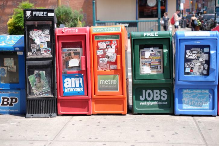 Aufgereihte Straßenzeitungsverkaufscontainer in unterschiedlichen Farben.