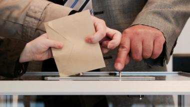 Jemand wirft einen braunen Briefumschkag i eine Wahlurne.