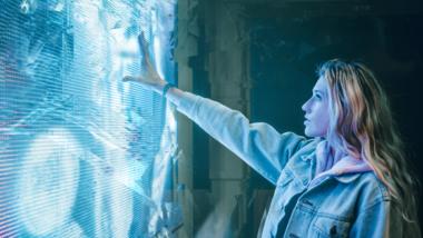 Eine junge Frau steht vor einer blau leuchtenden Kunstinstallation und streckt ihren Arm danach aus.