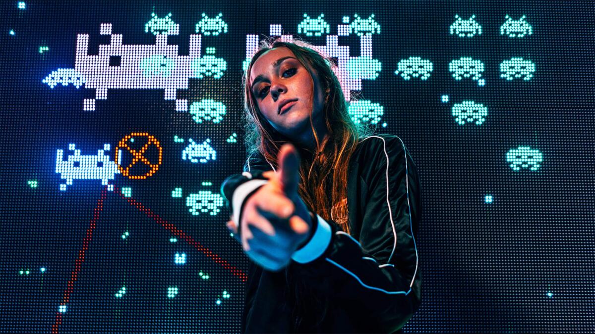 Frau deutet mit der Hand eine Pistole an, hinter ihr ein Bildschirm mit einem alten Computerspiel