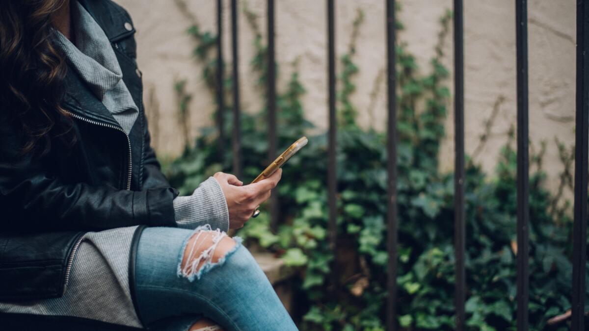 Frau sitzt mit Beinen übereinander geschlagen, i der Hand ein Smartphone