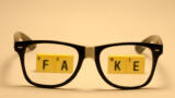 Eine Brille mit geklebtem Steg liegt auf einer orangenen Unterlage. Durch die Brillengläser sieht man das Wort FAKE, gelegt aus Scrabbel-Steinen.