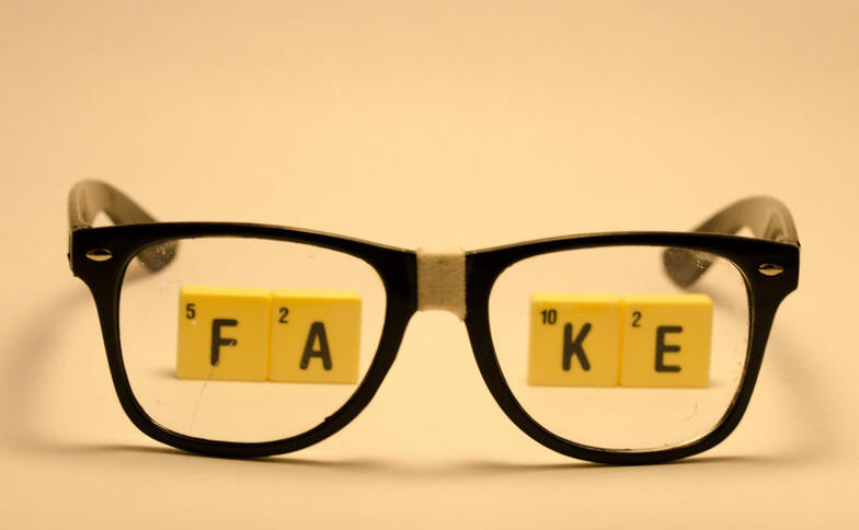 Eine Brille mit geklebtem Steg liegt auf einer orangenen Unterlage. Durch die Brillengläser sieht man das Wort FAKE, gelegt aus Scrabbel-Steinen.