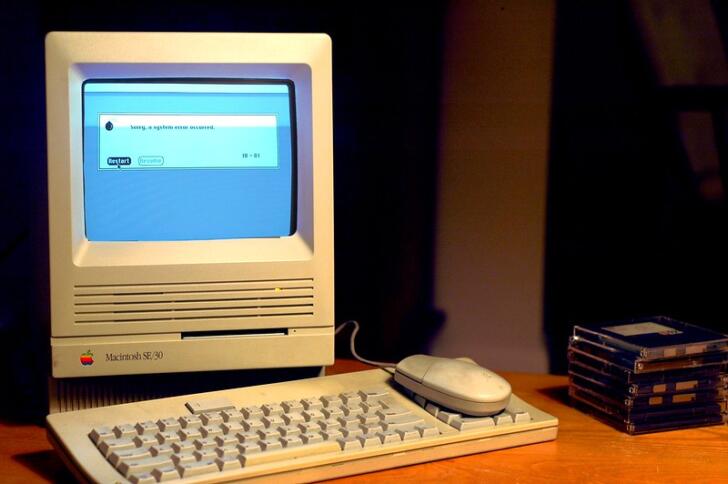 Man sieht ein sehr altes MacBook (vermutlich aus den Anfängen der Personal Comupter), das eine Fehlermeldung anzeigt, auf einem Schreibtisch.