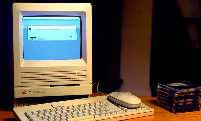 Man sieht ein sehr altes MacBook (vermutlich aus den Anfängen der Personal Comupter), das eine Fehlermeldung anzeigt, auf einem Schreibtisch.