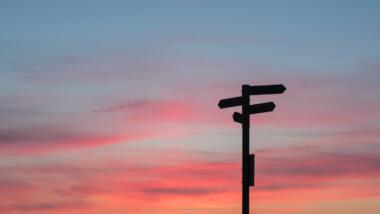 Ein Wegweiser mit vier Schildern, die in verschiedene Richtungen zeigen vor einem Abendhimmel ist zu sehen.