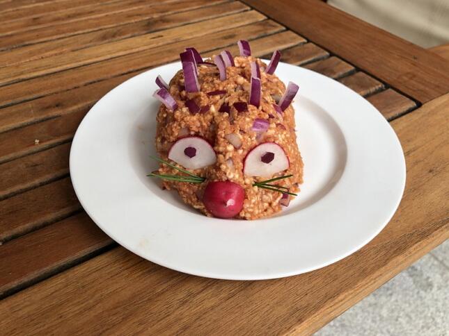 Auf einem Holztisch steht ein weißer Teller auf dem ein Mettigel angerichtet ist. Er hat eine Tomatennase.