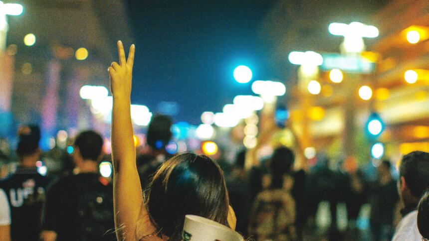 Man sieht eine Frau von hinten, sie ist auf einer Demonstration und hält die linke Hand hoch. Mit den Fingern macht sie das Victory-Zeichen.