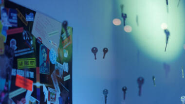 Die Ecke eines blau ausgeleuchteten Raumes ist zu sehen, an der linken Wand hängt eine Pinnwand mit vielen Stickern (einer davon ist ein "Asyl für Snowden"-Sticker). Davor scheinen Schlüssel in der Luft zu schweben, die an Schnüren an der Decke aufgehängt sind.