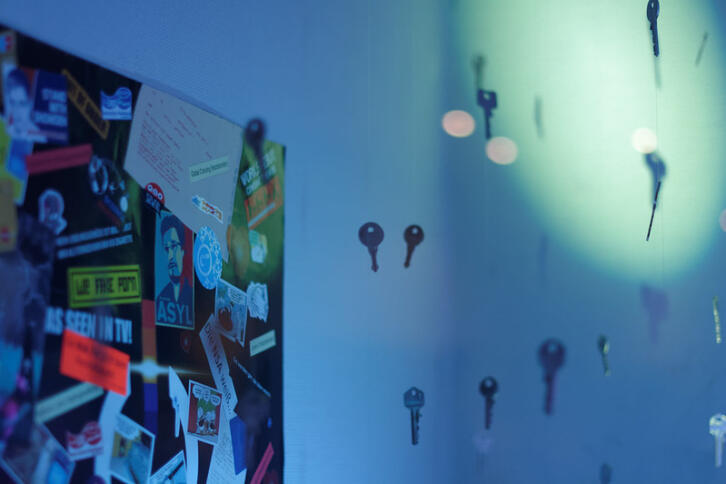 Die Ecke eines blau ausgeleuchteten Raumes ist zu sehen, an der linken Wand hängt eine Pinnwand mit vielen Stickern (einer davon ist ein "Asyl für Snowden"-Sticker). Davor scheinen Schlüssel in der Luft zu schweben, die an Schnüren an der Decke aufgehängt sind.