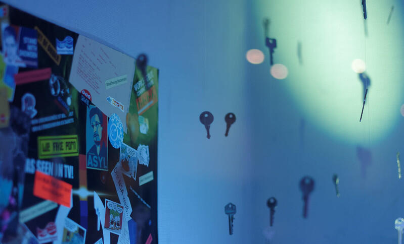 Die Ecke eines blau ausgeleuchteten Raumes ist zu sehen, an der linken Wand hängt eine Pinnwand mit vielen Stickern (einer davon ist ein "Asyl für Snowden"-Sticker). Davor scheinen Schlüssel in der Luft zu schweben, die an Schnüren an der Decke aufgehängt sind.