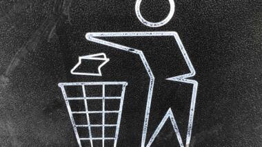 Man sieht ein Piktogramm auf schwarzem Hintergrund: Das Symbol für Wegwerfen beziehungsweise in den Müll tun.