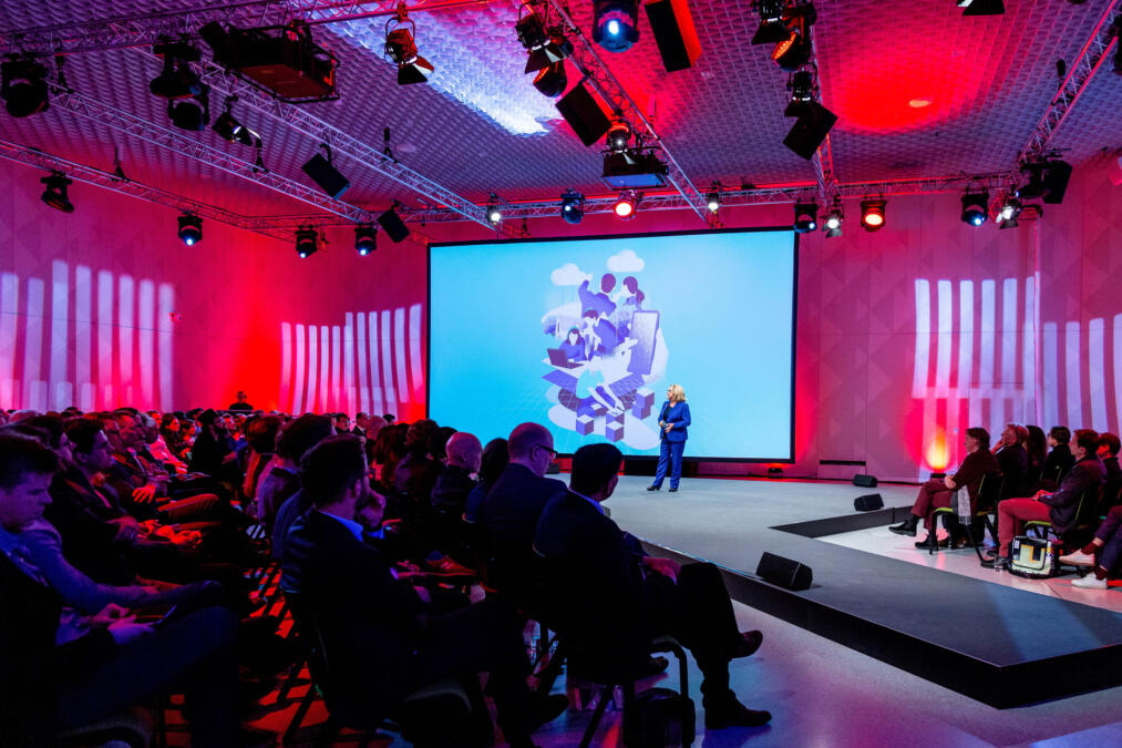 Ein großer Saal, in roten Farben beleuchtet. Zentral ist eine große Bühne, auf der die Ministerin Schulze vor einer Leinwand steht und eine Rede hält. Links und rechts davon sitzen Menschen in schicker Kleidung auf Stühlen im Publikum.