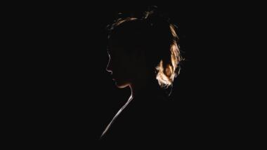 Die Silhouette einer Frau mit Pferdeschwanz-Frisur im Profil. Sie ist kaum zu erkennen, weil sie von hinten angeleuchtet wird. Lediglich ihre Umrisse heben sie vom ebenfalls schwarzen Hintergrund ab.