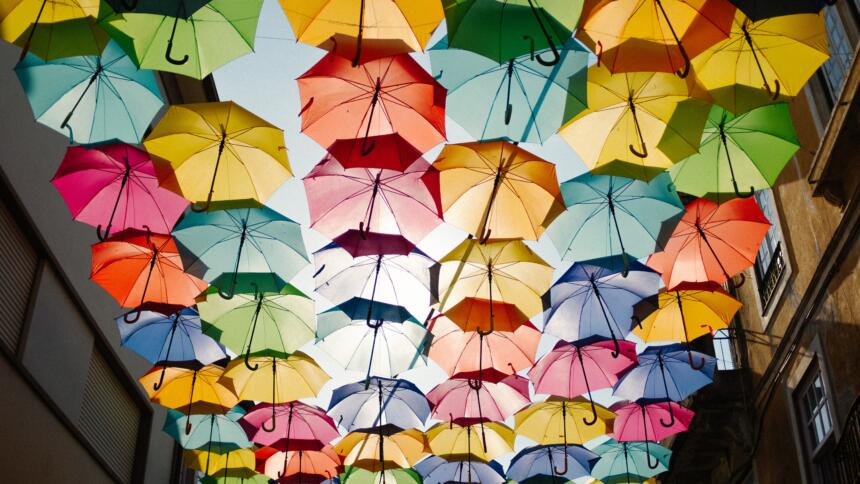 Eine Fotomontage: Von unten blickt man auf viele bunte Regenschirme, die zwischen zwei Häusern zu schweben scheinen und so quasi ein Dach bilden.