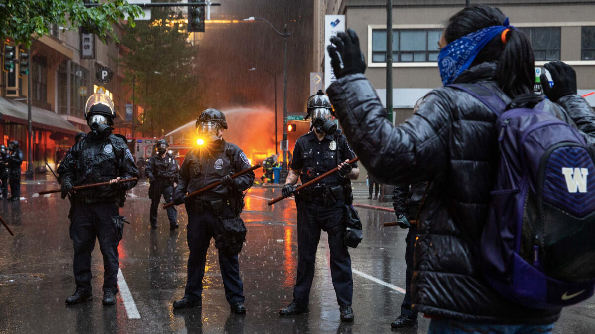 Frau steht mit erhobenen Armen vor einer Polizeikette, hinten löscht ein Feuerwehrfahrzeug einen Brand