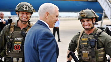 Mike Pence in blauem Jacket zwischen zwei Polizisten mit Helmen und schusssicheren Westen