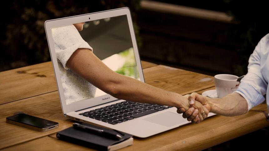 Aus einem Laptopbildschirm ragt ein menschlicher Arm, der einem realen Menschen, die Hand schüttelt.