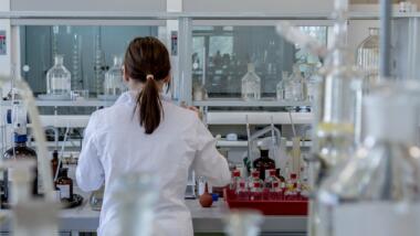 Frau steht im Labor umgeben von Kolben, Reagenzgläsern und anderen Gerätschaften.