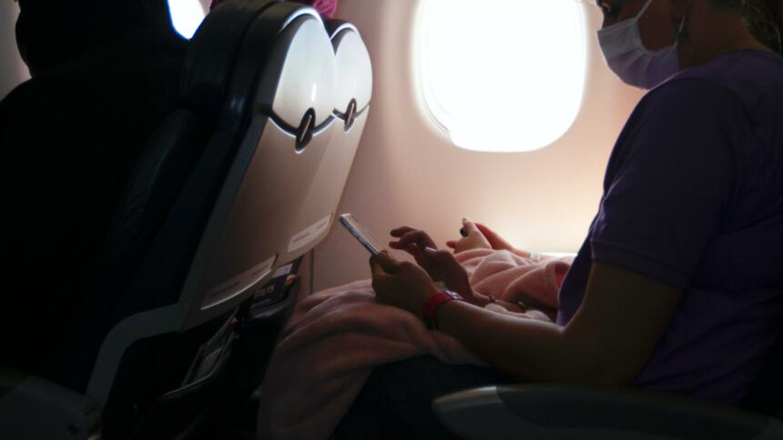 Person mit Maske sitzt in Flugzeug und tippt auf dem Smartphone