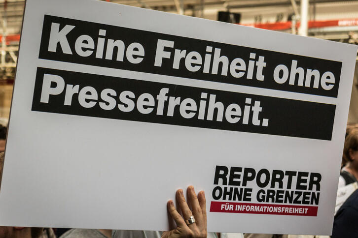 Hochgehaltenes weißes Schild auf dem in schwarz steht: "Keine Freiheit ohne Pressefreiheit" und ein Logo von Reporter ohne Grenzen zeigt.