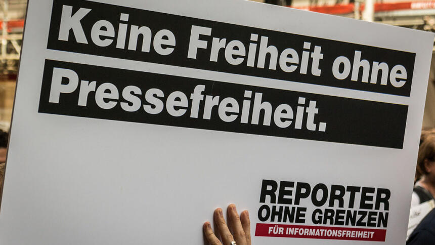 Hochgehaltenes weißes Schild auf dem in schwarz steht: "Keine Freiheit ohne Pressefreiheit" und ein Logo von Reporter ohne Grenzen zeigt.