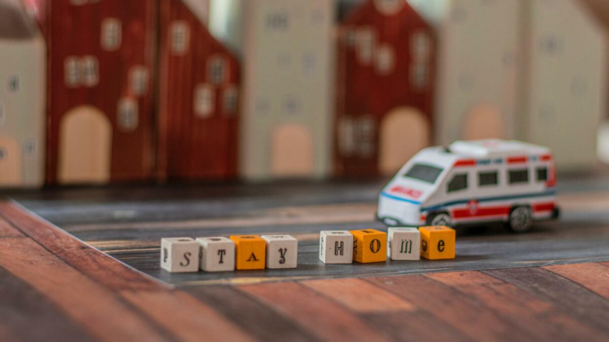 Spielzeug-Krankenwagen fährt an Häuser und dem Schriftzug "Stay home" auf Bauklötzchen vorbei.