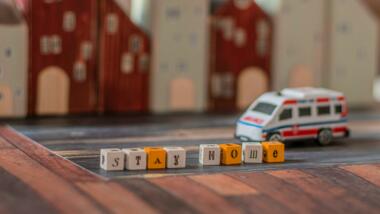 Spielzeug-Krankenwagen fährt an Häuser und dem Schriftzug "Stay home" auf Bauklötzchen vorbei.