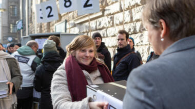 Eine Frau übergibt vor einem großen Gebäude in Berlin mehrere Aktenordner.