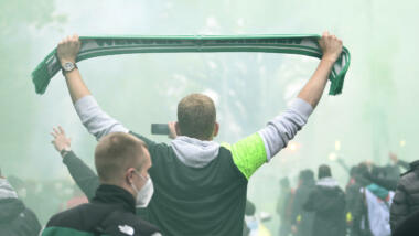 Fan von Werder Bremen steht zwischen anderen grüngekleideten Fans und hält grünen Fanschal hoch. Im Hintergrund grüne Rauchschwaden.