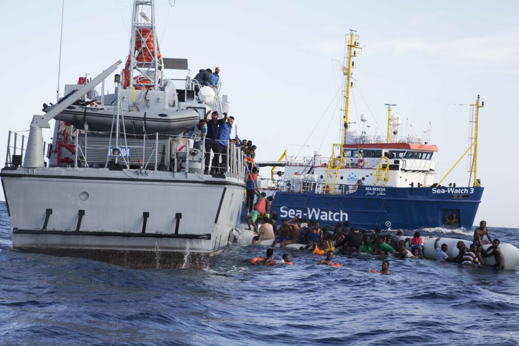 Das Bild zeigt Geflüchtete im Wasser vor einem libyschen Küstenwachschiff, im Hintergrund ein Schiff von Sea-Watch.
