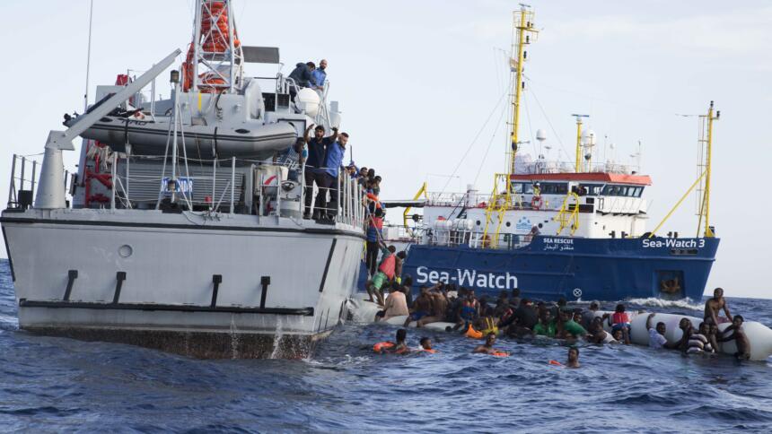 Das Bild zeigt Geflüchtete im Wasser vor einem libyschen Küstenwachschiff, im Hintergrund ein Schiff von Sea-Watch.