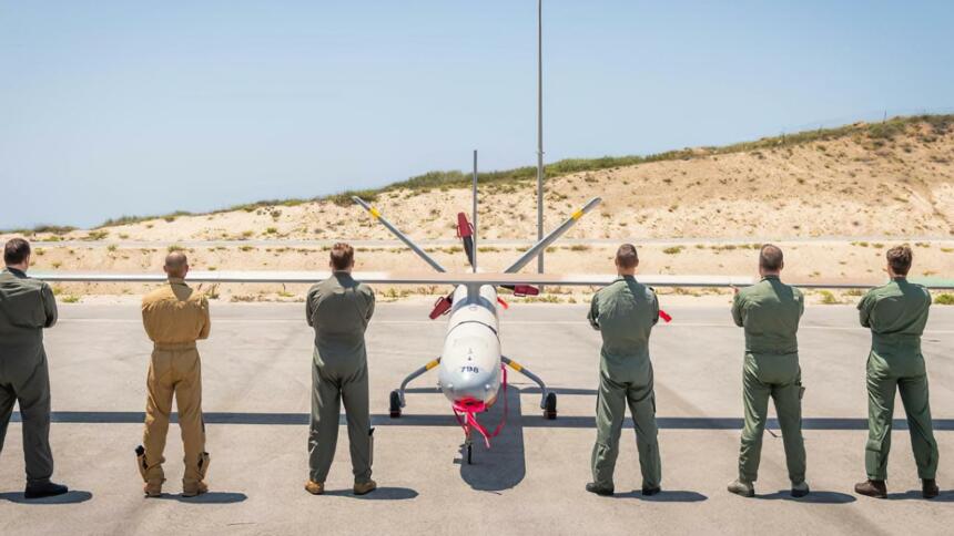 Das Bild zeigt eine Drohne, vor der sechs Soldat:innen mit verschränkten Armen und mit dem Rücken zur Kamera stehen.