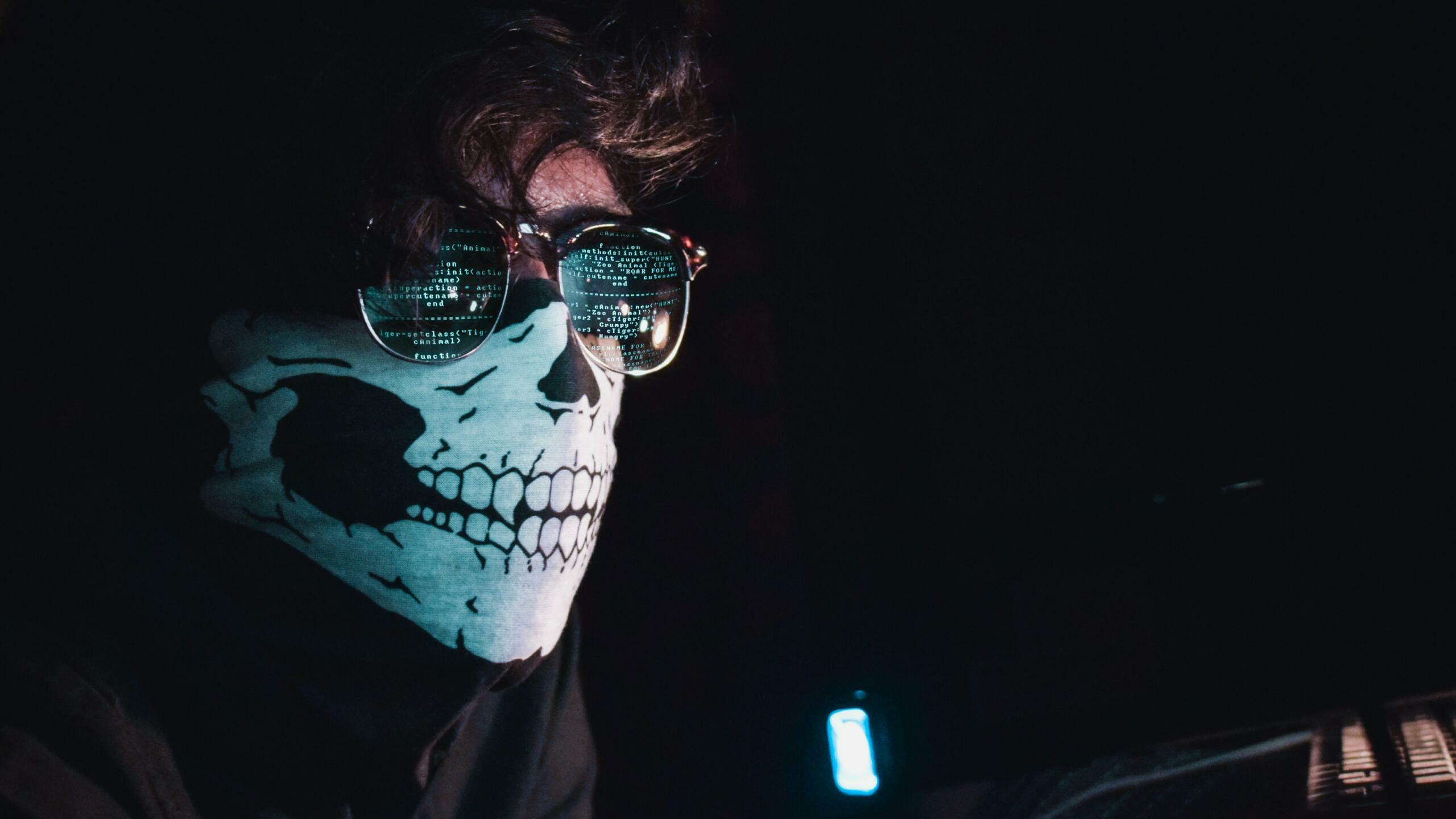 Mann mit Totenkopfmaske und Sonnebrille sitzt in dunklem Raum.