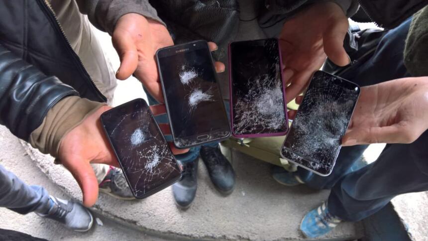 Das Bild zeigt die Hände von vier Personen, die Mobiltelefone mit zerstörten Displays in der Hand halten.