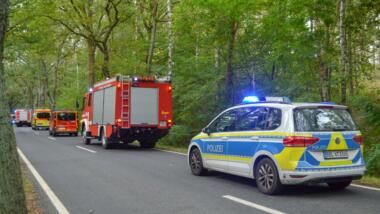 Straße durch einen Wald, befahren von Fahrzeugen der Polizei, der Feuerwehr und des Rettungsdienstes, teilweise mit Blaulicht.