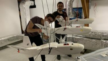 Das Bild zeigt zwei Männer, die an vier Drohnen arbeiten die an Schnüren hängen.