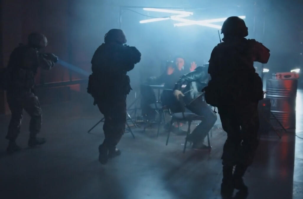 Das Bild zeigt die von hinten Silhouetten bewaffneter Spezialeinheiten, die sich im Dunlen auf einen belechteten Tisch zubewegen an dem zwei junge Männer vor Bildschirmen sitzen.