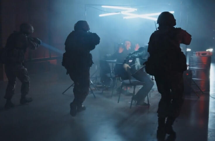 Das Bild zeigt die von hinten Silhouetten bewaffneter Spezialeinheiten, die sich im Dunlen auf einen belechteten Tisch zubewegen an dem zwei junge Männer vor Bildschirmen sitzen.