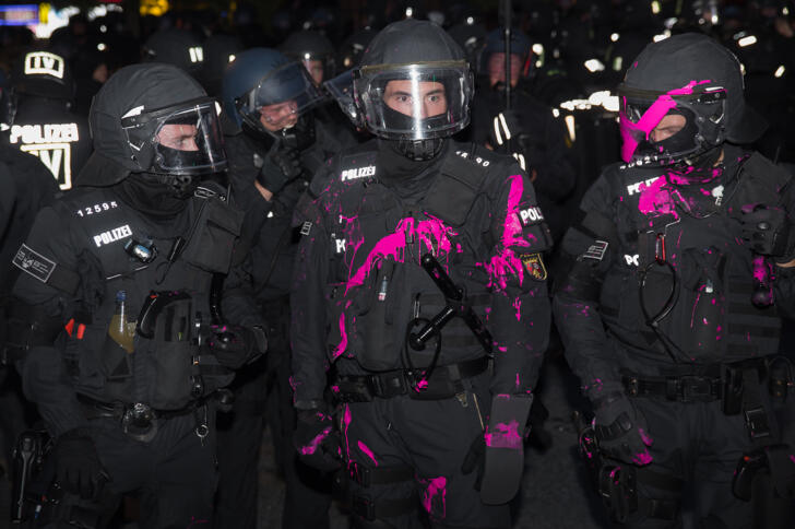 Das Bild zeigt mehrere Polizisten in schwarzen Uniformen im Dunkeln, von denen zwei mit Farbe in Pink bekleckert sind.