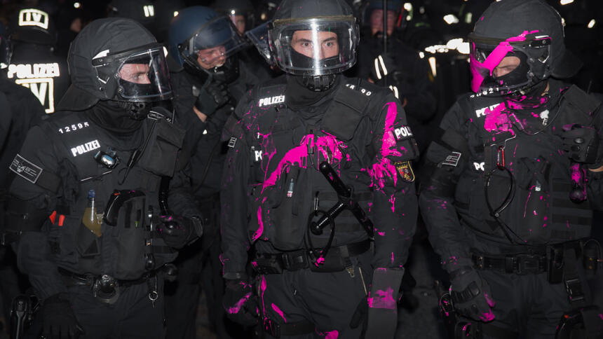 Das Bild zeigt mehrere Polizisten in schwarzen Uniformen im Dunkeln, von denen zwei mit Farbe in Pink bekleckert sind.
