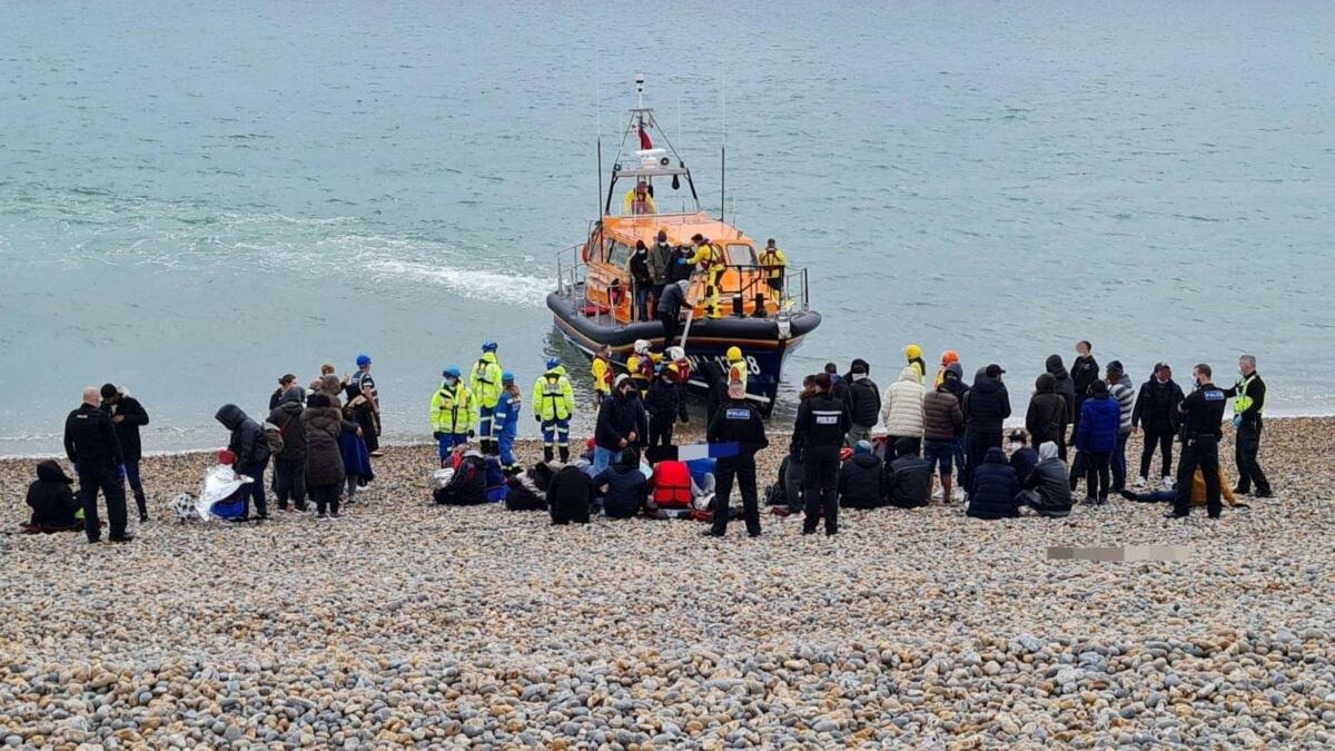 Das Bild zeiht einen kiesigen Strand, an dem viele, teils uniformierte Menschen um einige am Boden sitzende Menschen herumstehen, im Wasser liegt ein Seenotrettungsboot mit Kajüte.