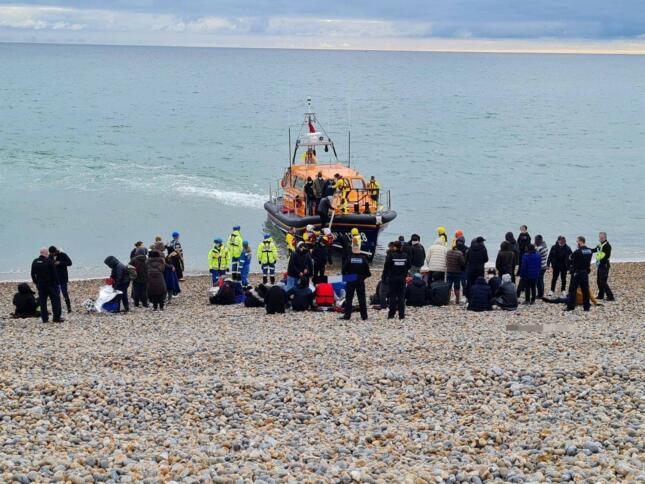 Das Bild zeiht einen kiesigen Strand, an dem viele, teils uniformierte Menschen um einige am Boden sitzende Menschen herumstehen, im Wasser liegt ein Seenotrettungsboot mit Kajüte.