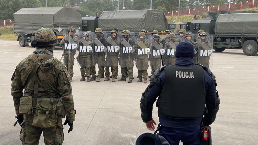 Das Bild zeigt eine Gruppe von Militärs mit Helmen und Schilden, im Hintergrund sindmehrere olivgrüne Lastkraftwagen zu sehen. Im Vordergrund beobachten ein Soldat und ein Polizist das Geschehen.