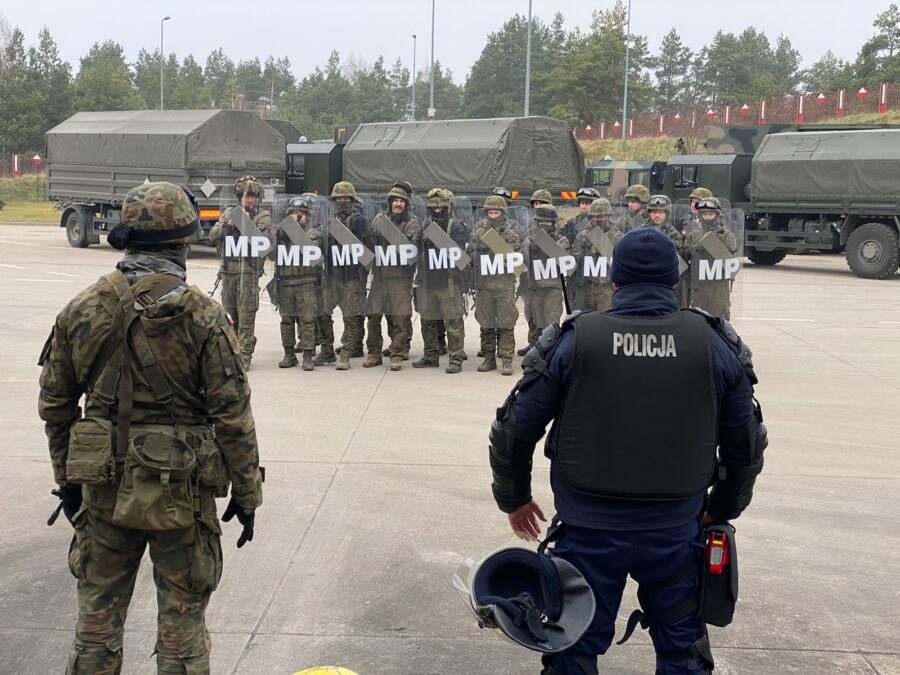 Das Bild zeigt eine Gruppe von Militärs mit Helmen und Schilden, im Hintergrund sindmehrere olivgrüne Lastkraftwagen zu sehen. Im Vordergrund beobachten ein Soldat und ein Polizist das Geschehen.
