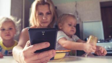Mutter mit zwei Kleinkindern auf dem Schoss hält ein Smartphone in der Hand.